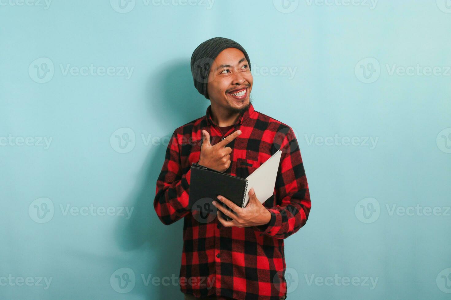 An excited young Asian man with a beanie hat and a red plaid flannel shirt is pointing his finger aside the copyspace while holding a book, standing against a blue background photo