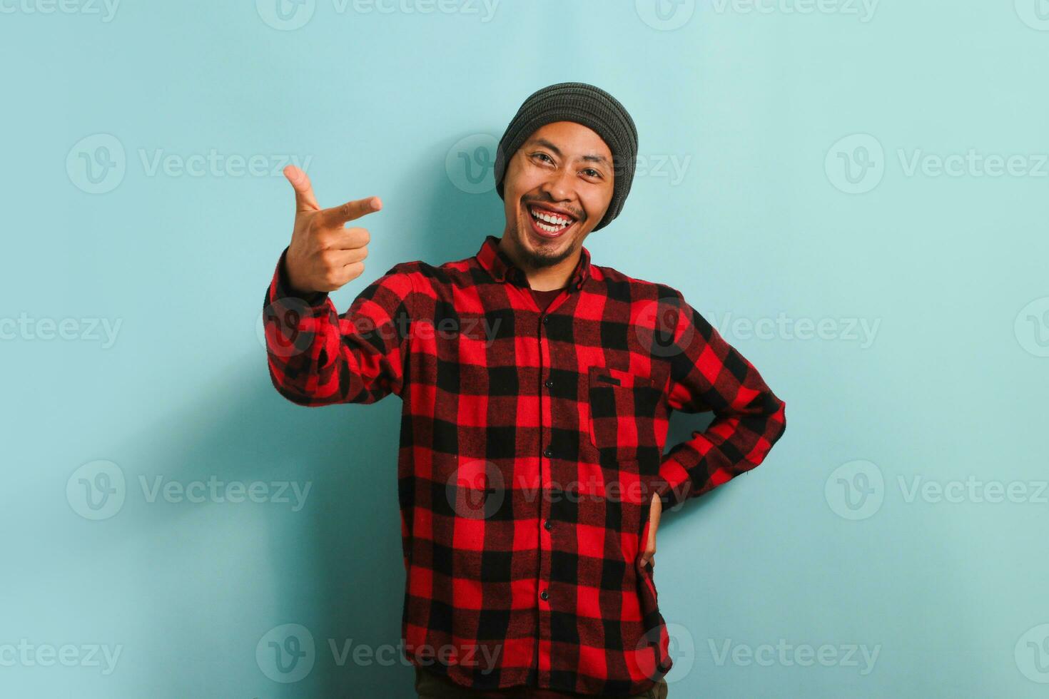 Confident young Asian man with a beanie hat and a red plaid flannel shirt is pointing his finger at the camera and smiling, choosing, inviting people, or recruiting. He is isolated on blue background photo