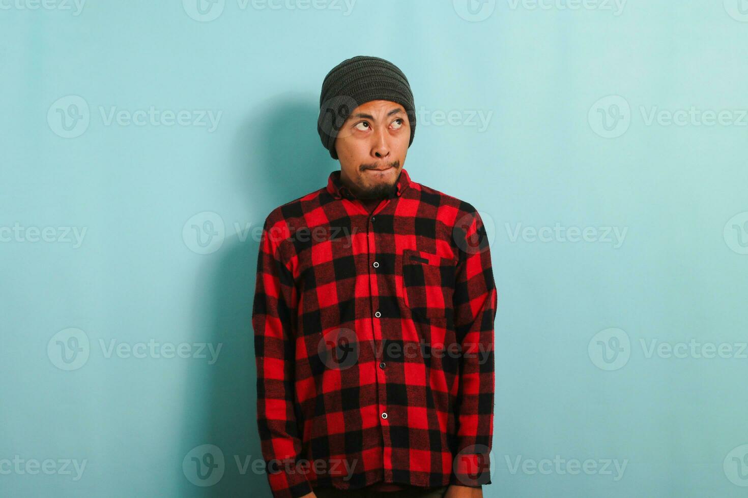 A confused young Asian man with a beanie hat and a red plaid flannel shirt pursing his lips as if forbidden to say anything while standing against a blue background photo