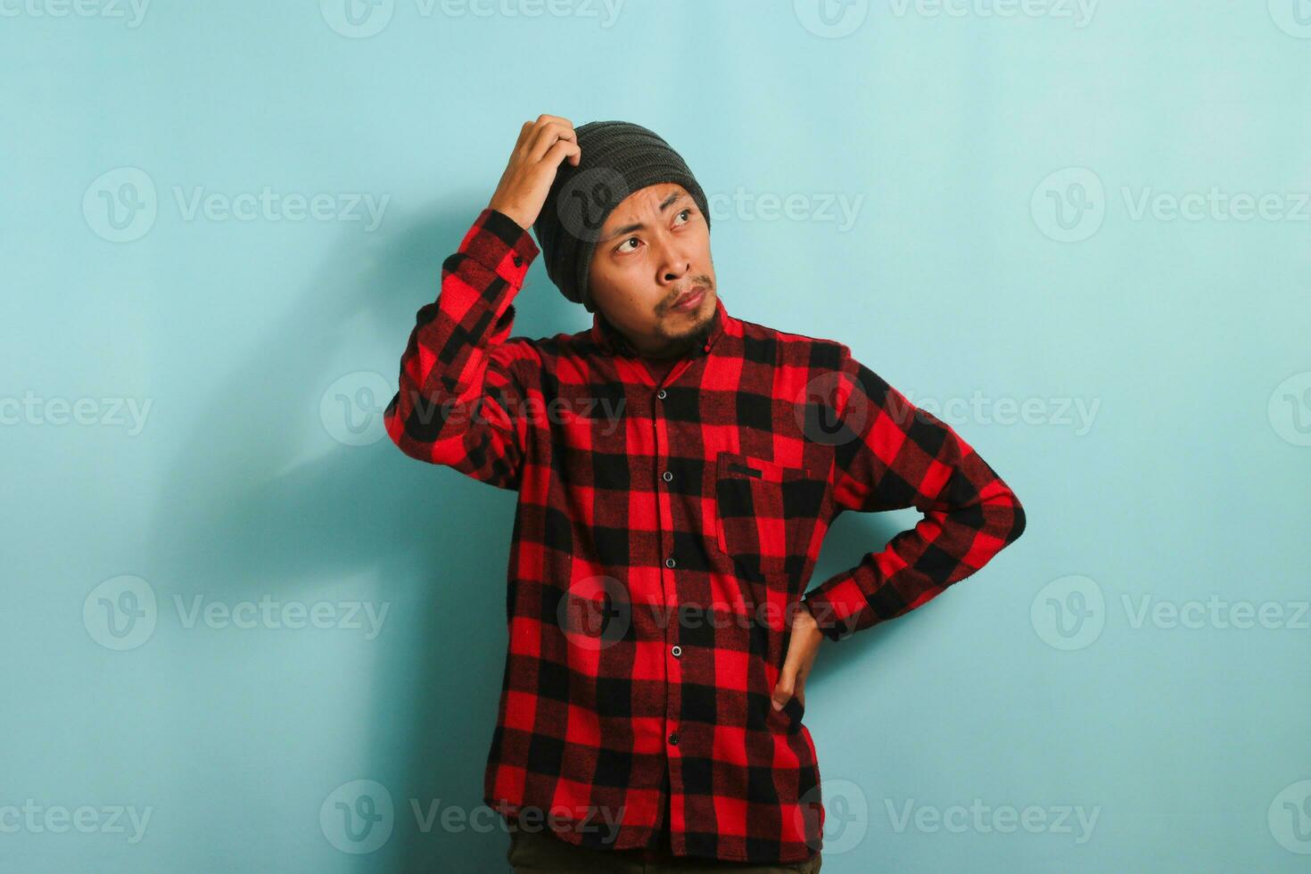 A confused young Asian man with a beanie hat and a red plaid flannel shirt scratching his head and looking up at the copy space while standing against a blue background photo