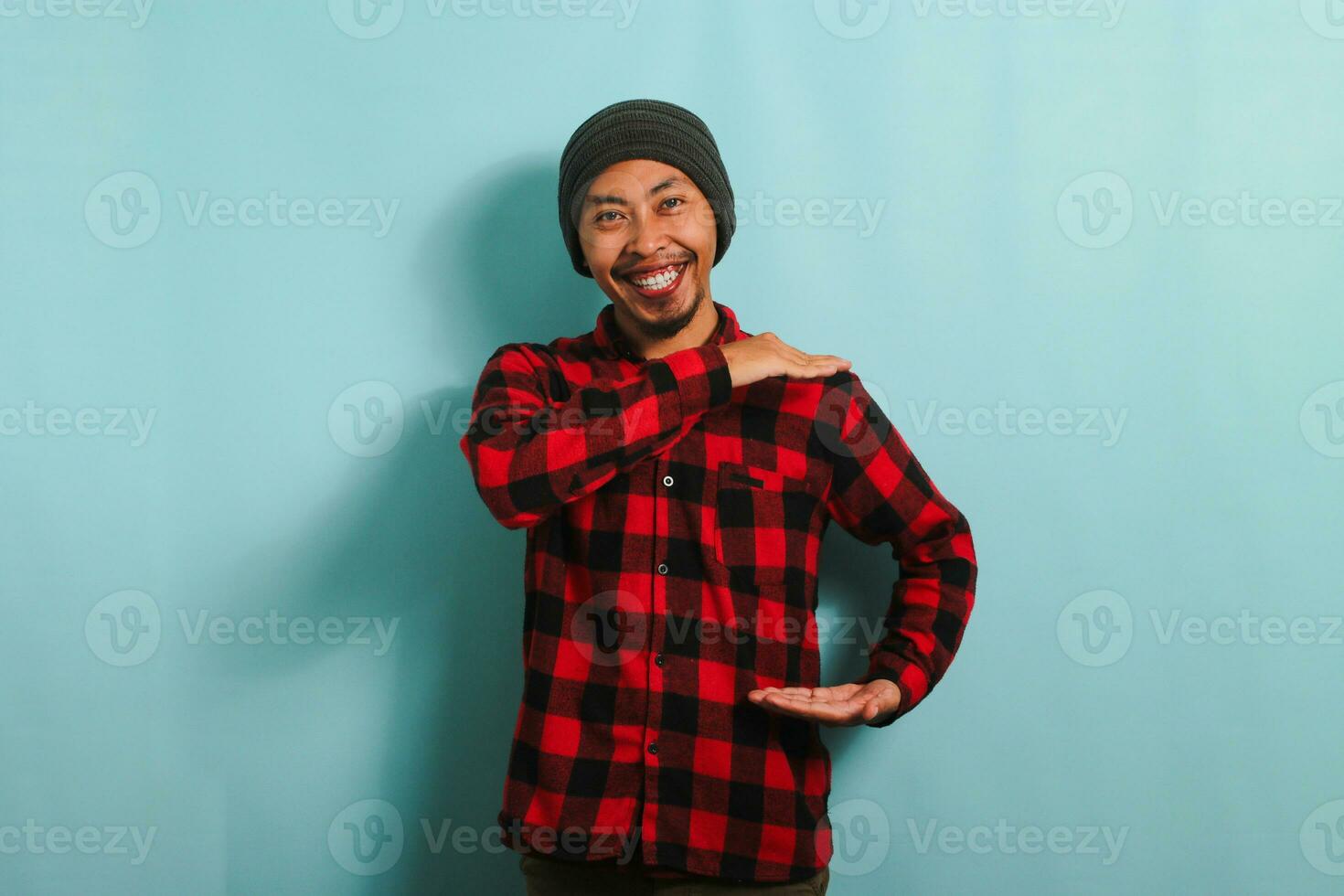 Young Asian man with beanie hat and red plaid flannel shirt gesturing a sign of size with both hands, measuring something, isolated on a blue background photo