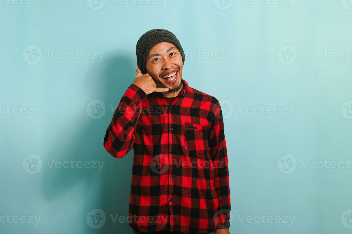An excited young Asian man wearing a beanie hat and a red plaid flannel shirt makes a CALL ME gesture with his hand, isolated on a blue background photo