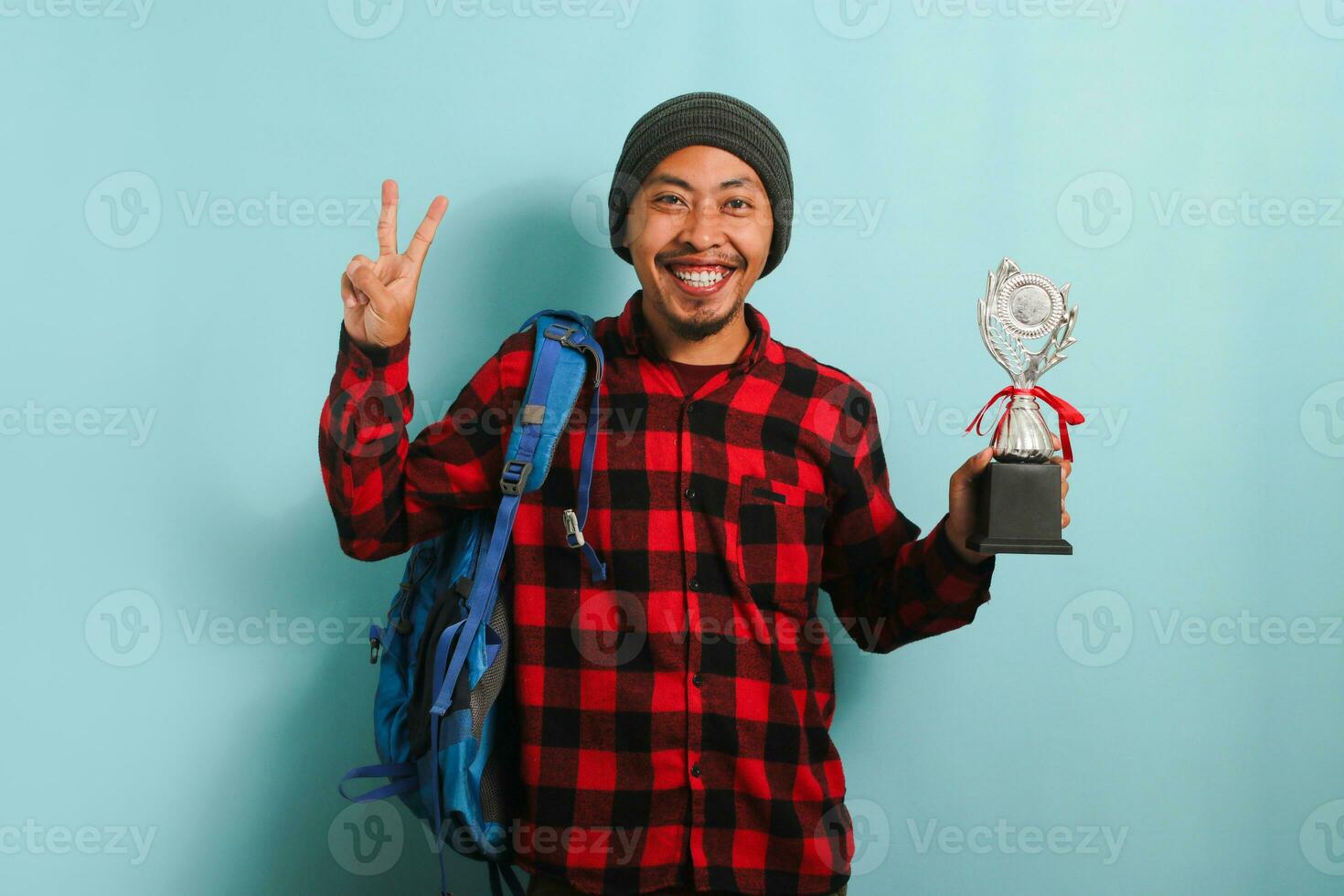 Excited young Asian man student wearing a backpack, beanie hat, and red plaid flannel shirt, making a victory gesture while holding a trophy and rejoicing in achievement, isolated on a blue background photo