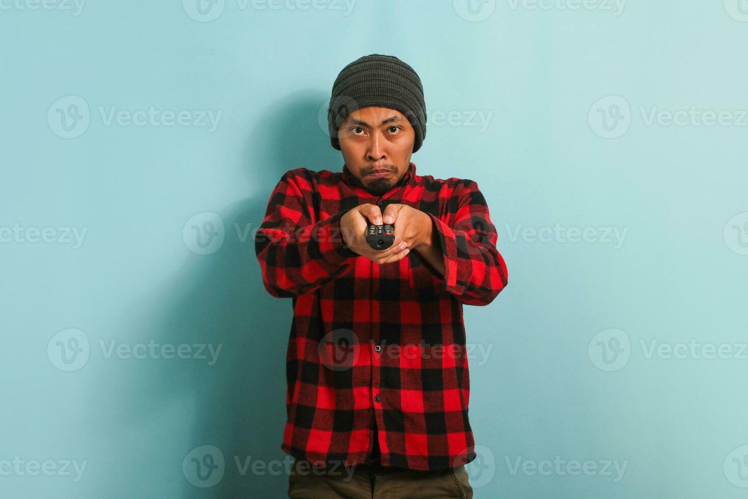 An annoyed young Asian man with a beanie hat and red plaid flannel shirt is switching TV channels using the TV remote, feeling upset and bored, isolated on a blue background. photo