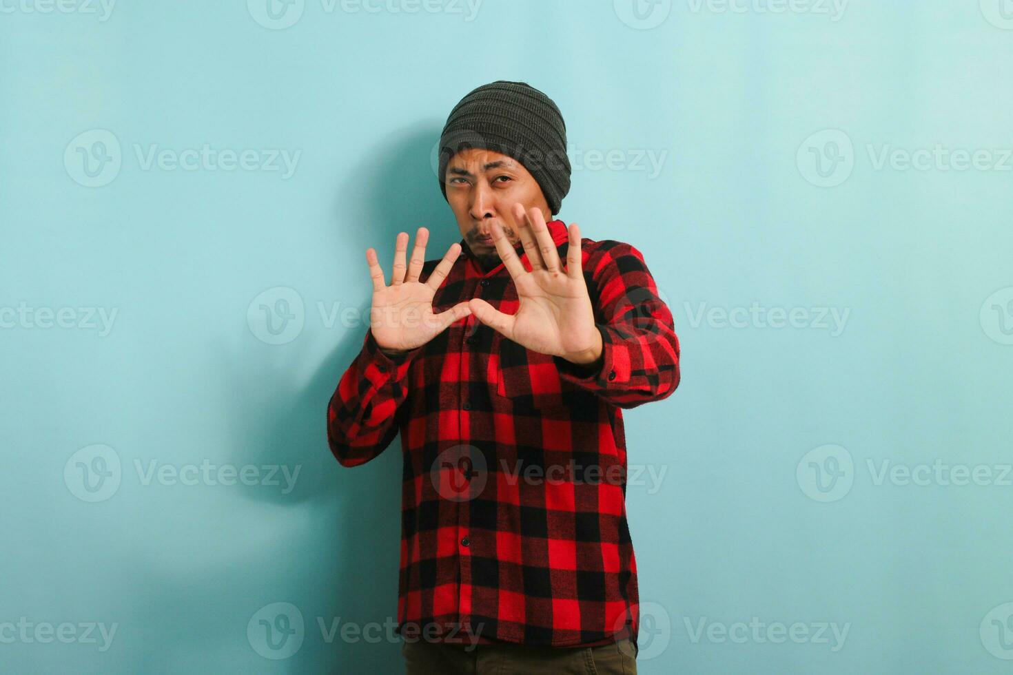 Ew, It's so gross. An annoyed young Asian man with a beanie hat and a red plaid flannel shirt is repulsed by something, displaying a disgusted expression while standing against a blue background photo