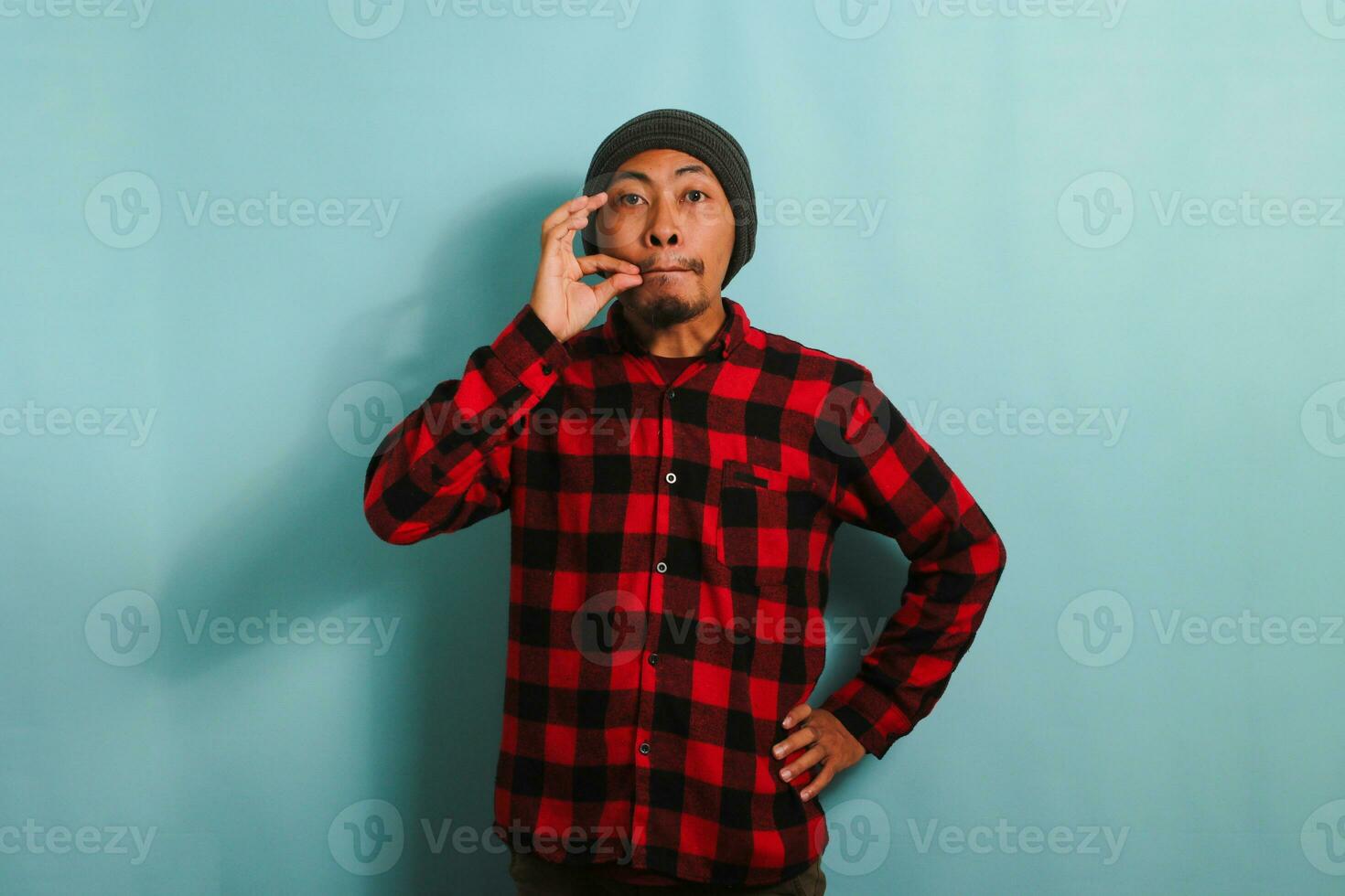 Young Asian man with beanie hat and red plaid flannel shirt makes a zipping and locking mouth shut gesture, isolated on a blue background. Keeping Secret, silent, taboo talking concept. photo