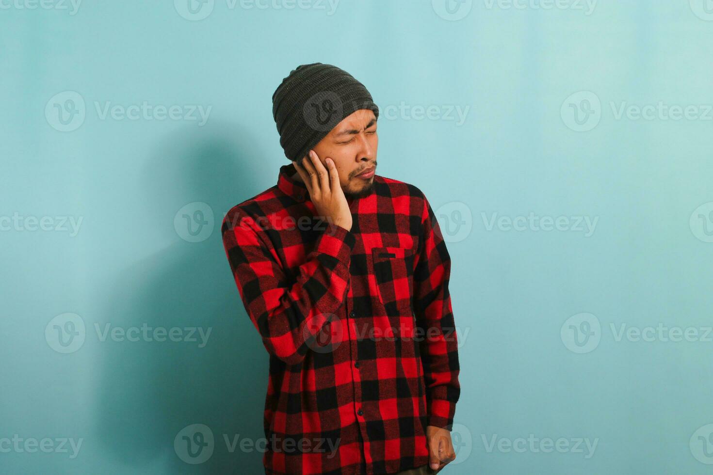 Young Asian man with beanie hat and red plaid flannel shirt holds his hand near his ear, suffering from tinnitus, a throbbing earache, and fatigue due to noise, while standing against blue background photo