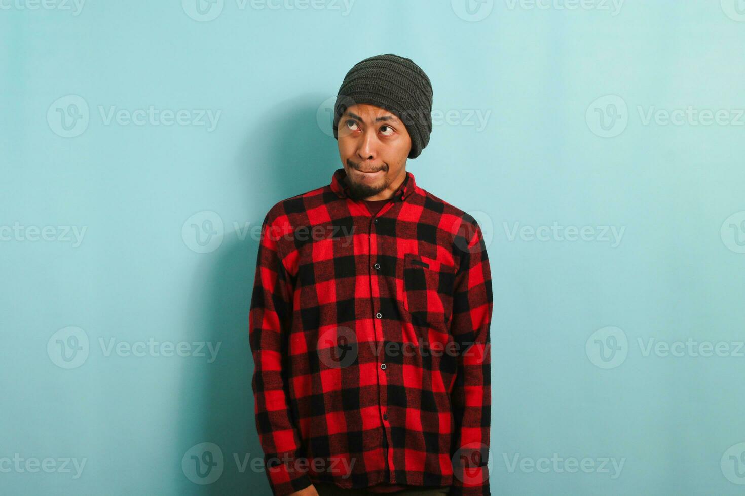 A confused young Asian man with a beanie hat and a red plaid flannel shirt pursing his lips as if forbidden to say anything while standing against a blue background photo