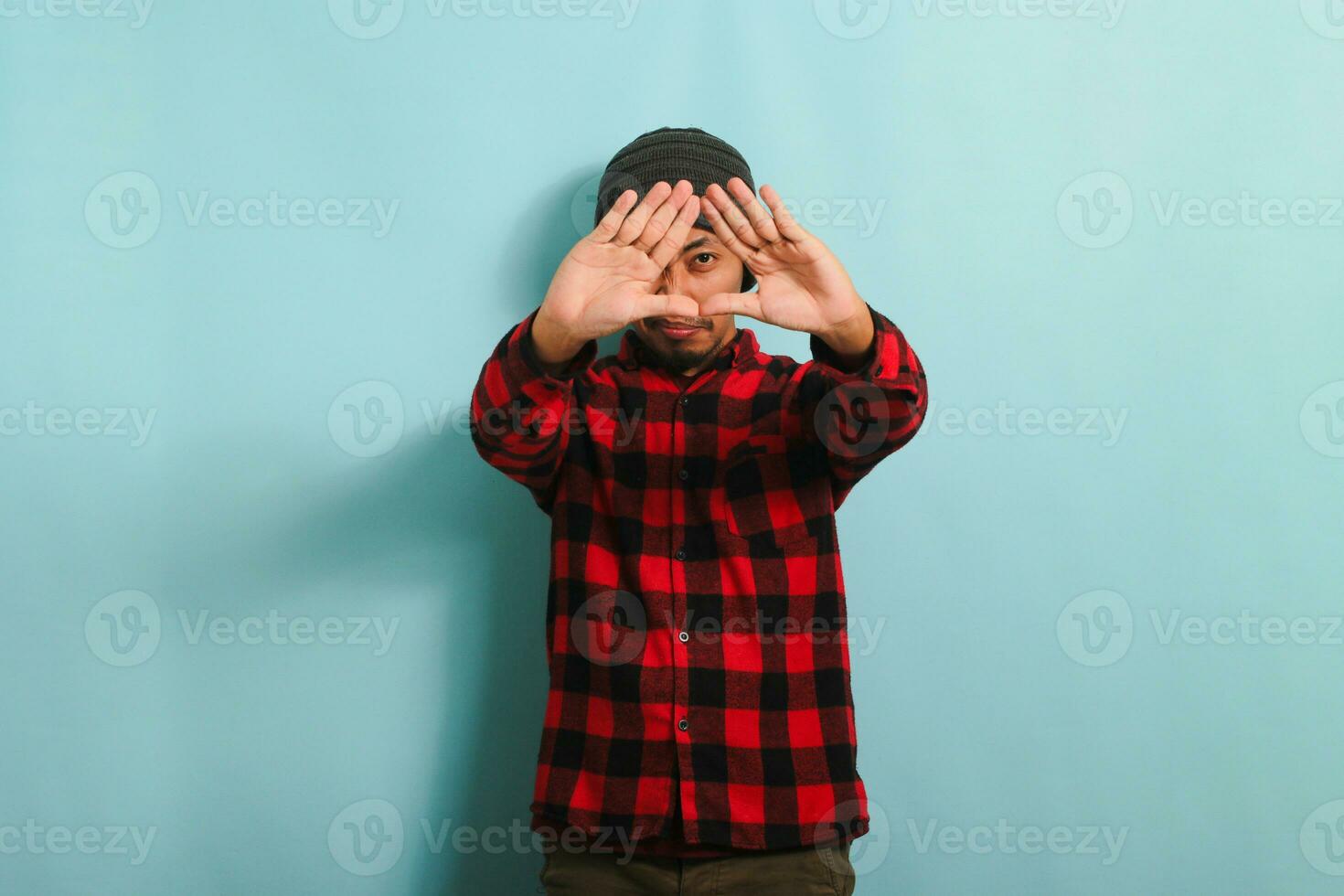 Young Asian man with beanie hat and red plaid flannel shirt making a triangle gesture with palms, symbolizing the environment protection, recycling, and reusing concept, isolated on a blue background photo