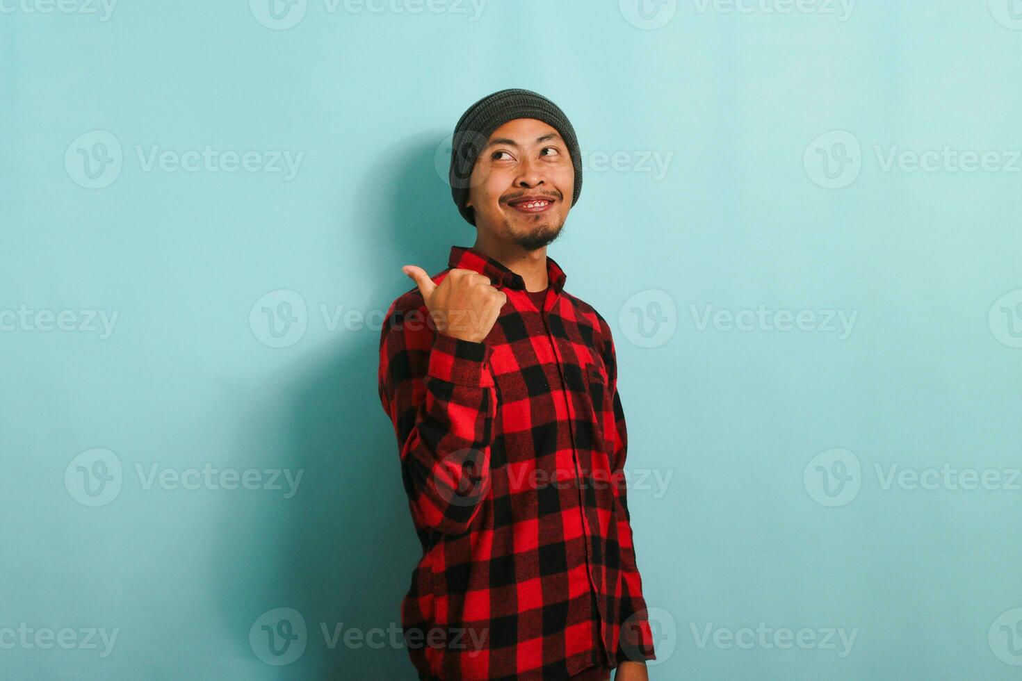 Attractive Young Asian man with beanie hat and red plaid flannel shirt pointing with his thumb at empty copy space, isolated on a blue background photo