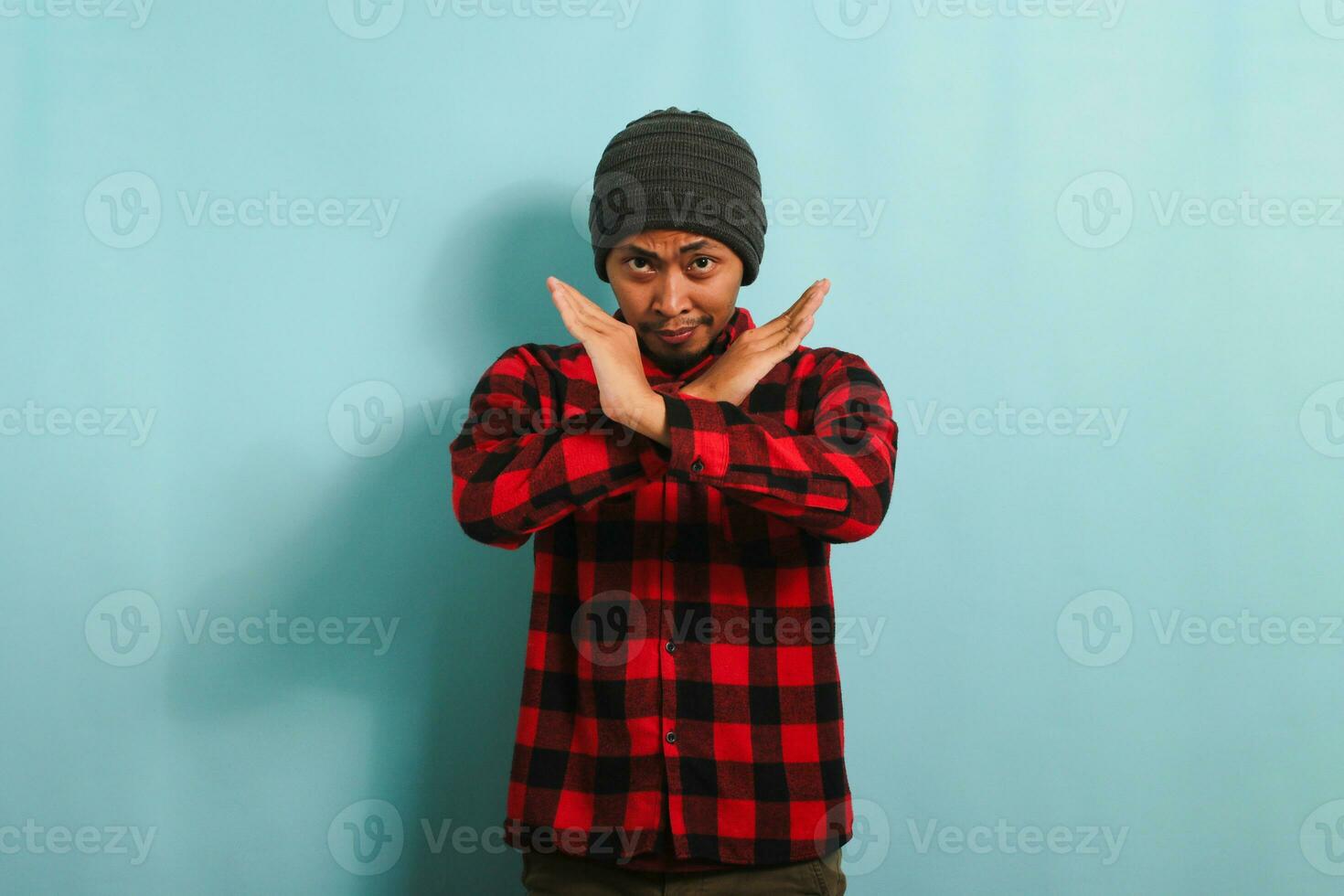 An angry young Asian man with a beanie hat and a red plaid flannel shirt stands with his arms crossed, displaying a refusal, denial, and stop gesture while standing against a blue background. photo