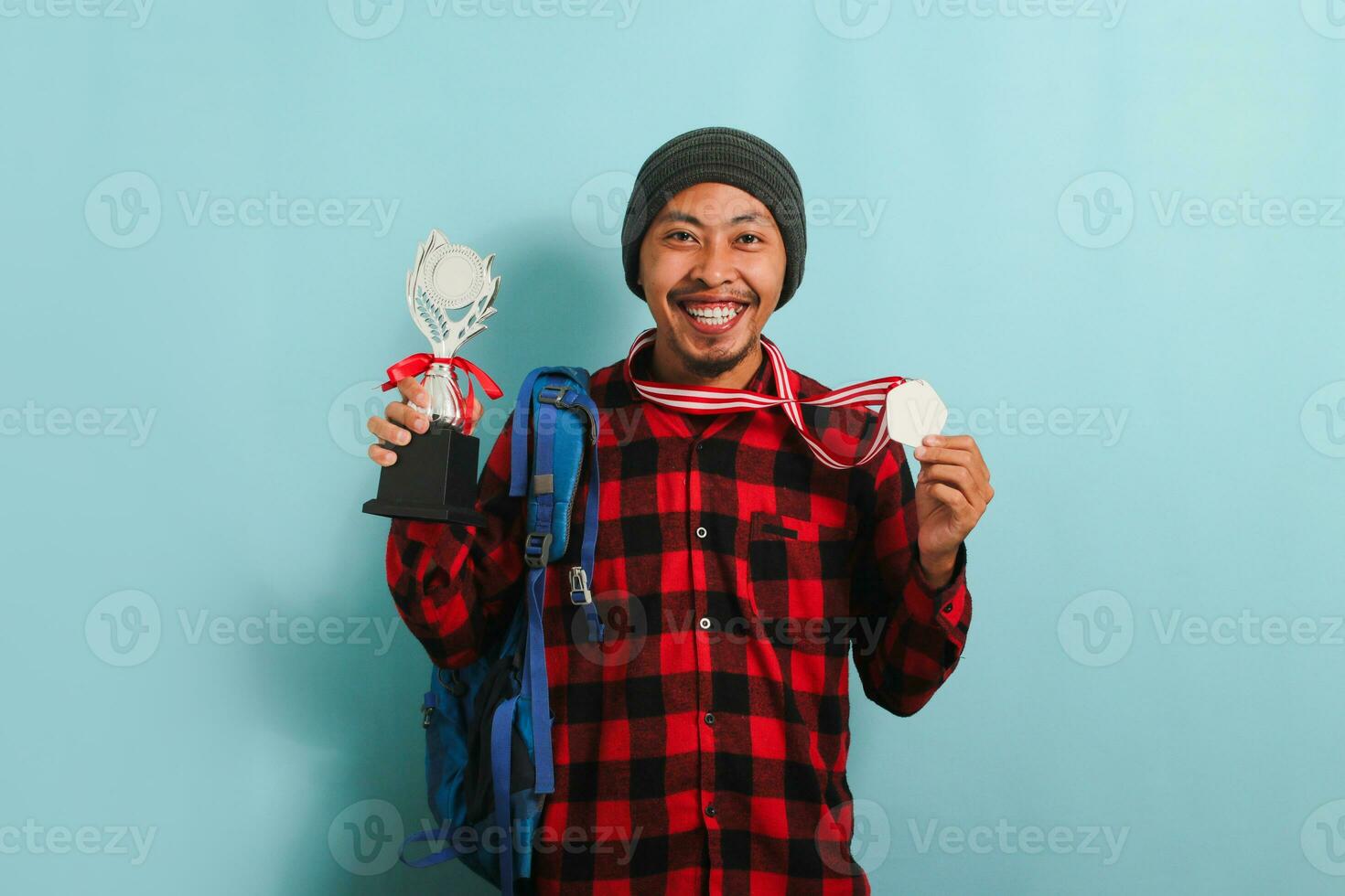 Happy young Asian man student wearing a backpack, beanie hat, and red plaid flannel shirt, showing a medal and trophy while rejoicing in success and achievement, isolated on a blue background photo