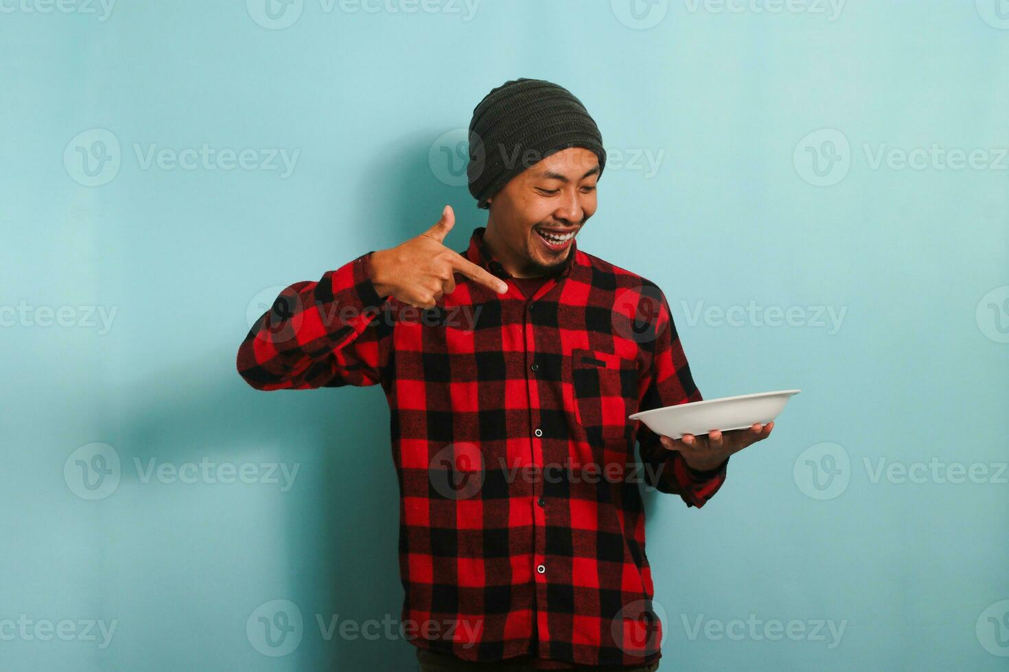 Excited Young Asian man with a beanie hat and a red plaid flannel shirt is pointing to an empty white plate in his hand, isolated on a blue background photo