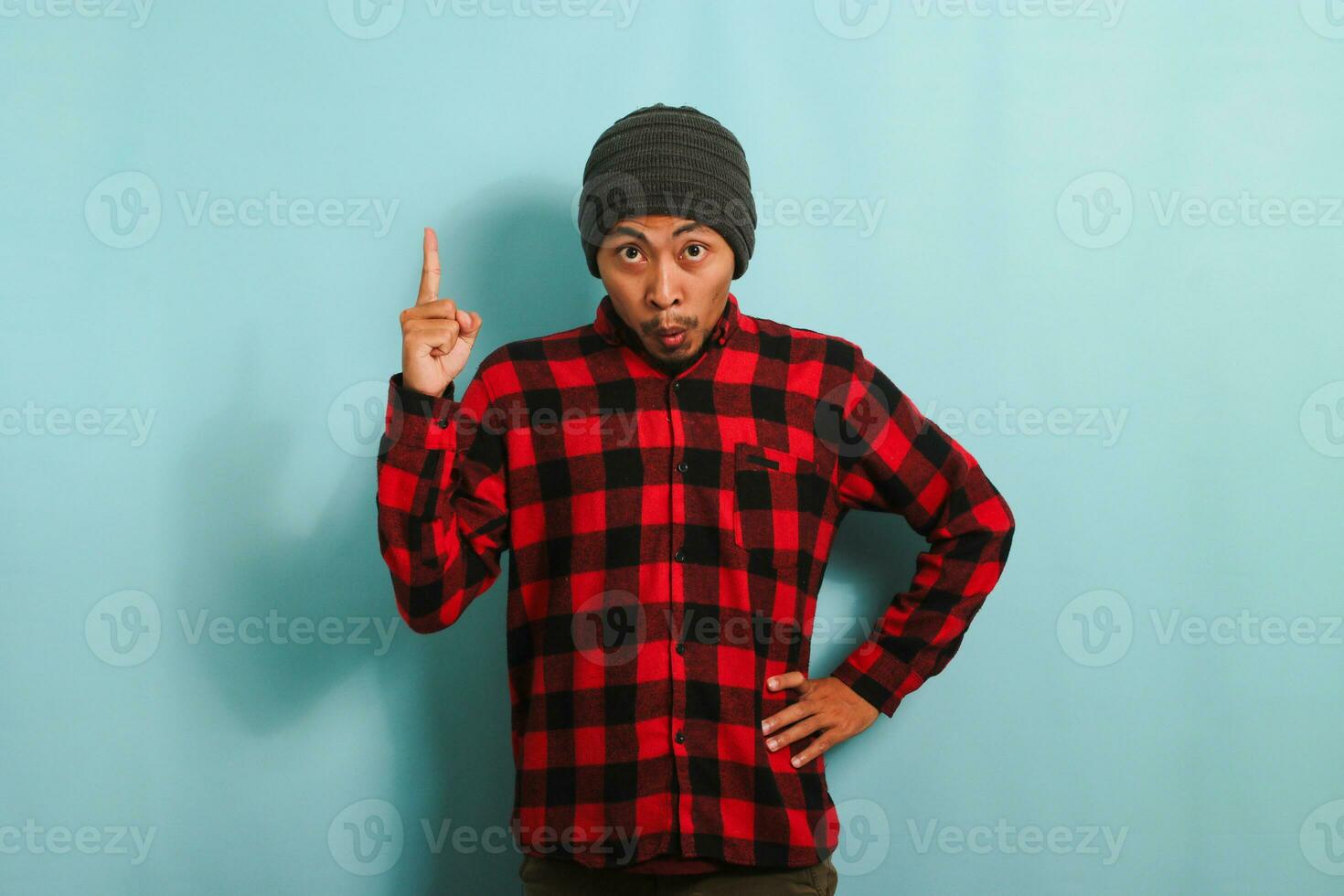 Surprised Young Asian man with a beanie hat and red plaid flannel shirt is pointing his finger up towards the copy space, isolated on a blue background photo