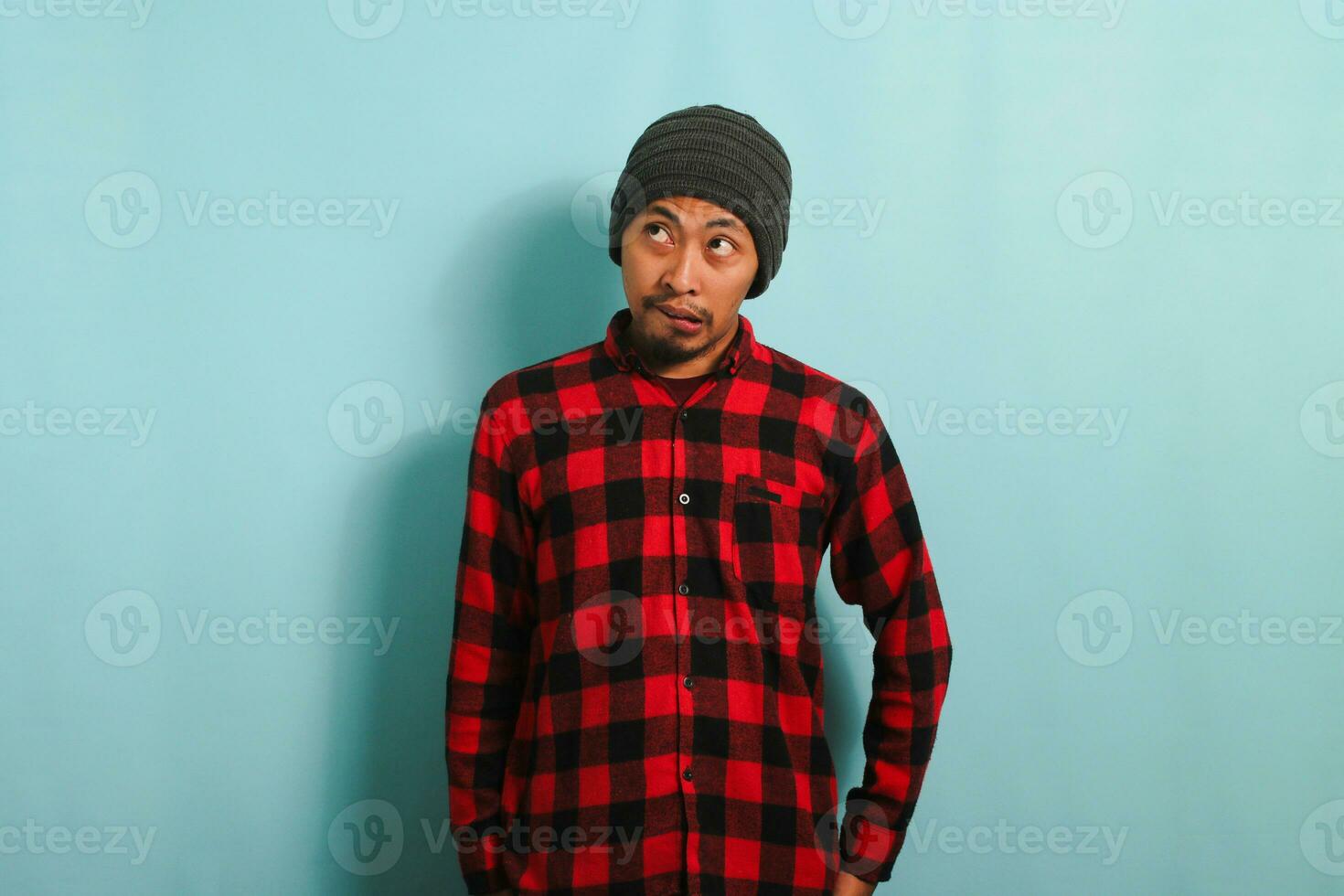 An embarrassed young Asian man with a beanie hat and a red plaid flannel shirt bites his lower lip while looking aside at an empty copyspace, while standing against a blue background photo