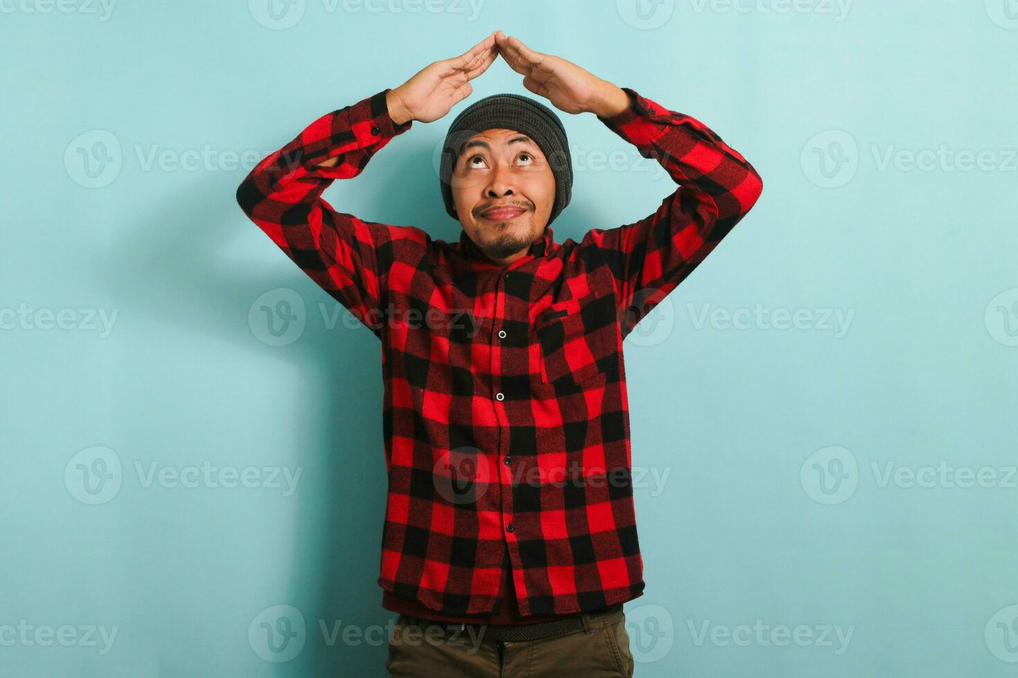 Happy young Asian man with a beanie hat and red plaid flannel shirt making a roof gesture above his head with a beaming smile, isolated on a blue background photo