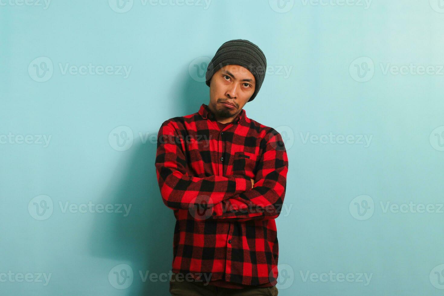 An angry young Asian student man wearing a beanie hat and a red plaid flannel shirt frowns and keeps his arms folded, appearing bored, isolated on a blue background photo