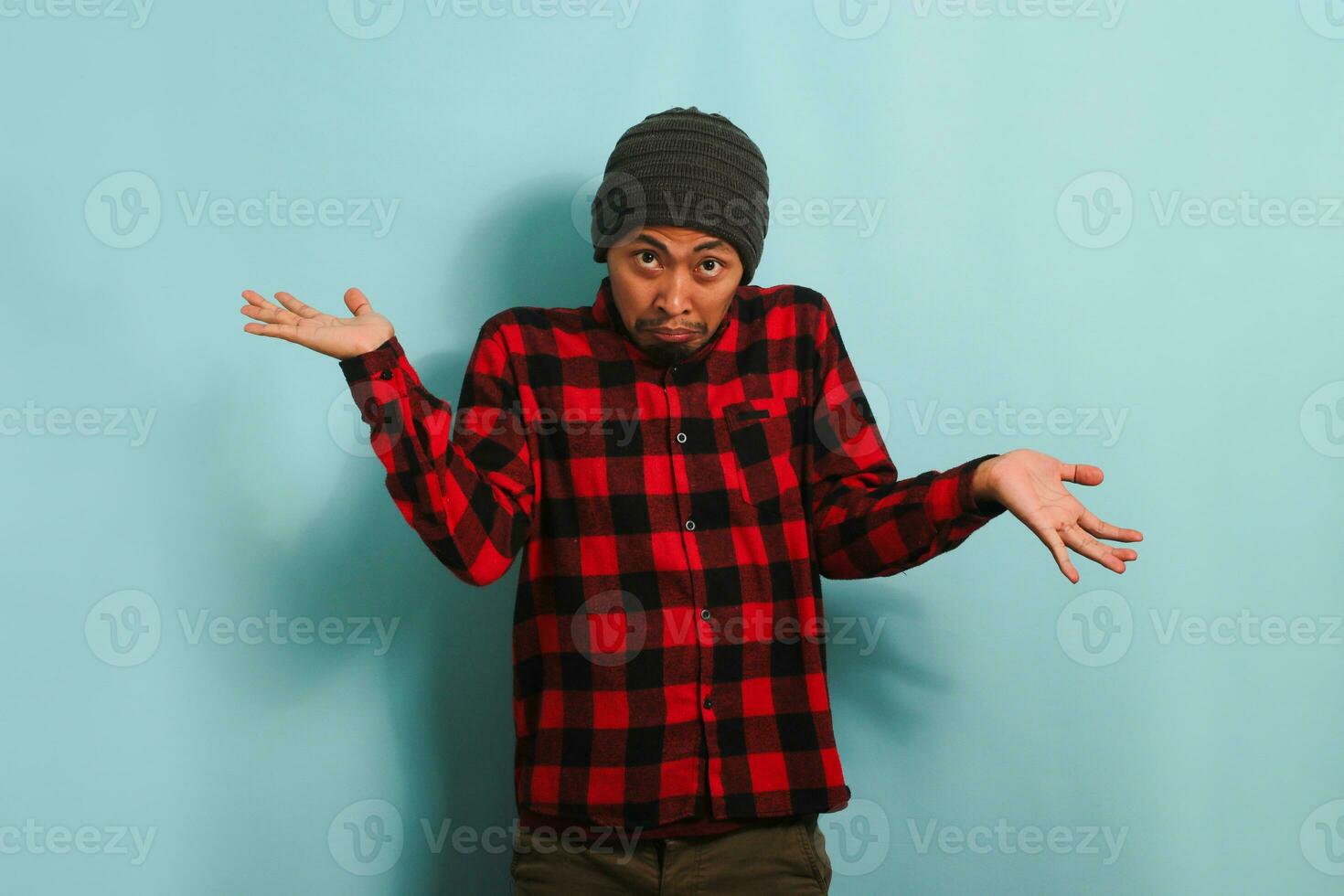 A confused young Asian man with a beanie hat and a red plaid flannel shirt is shrugging his shoulders and looking at the camera with a puzzled expression, isolated on a blue background. photo