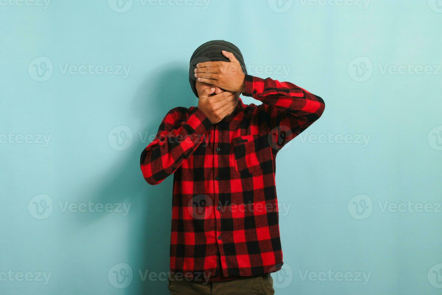 Depressed young Asian man with a beanie hat and a red plaid flannel shirt covering his face with his hands, crying while standing against a blue background photo