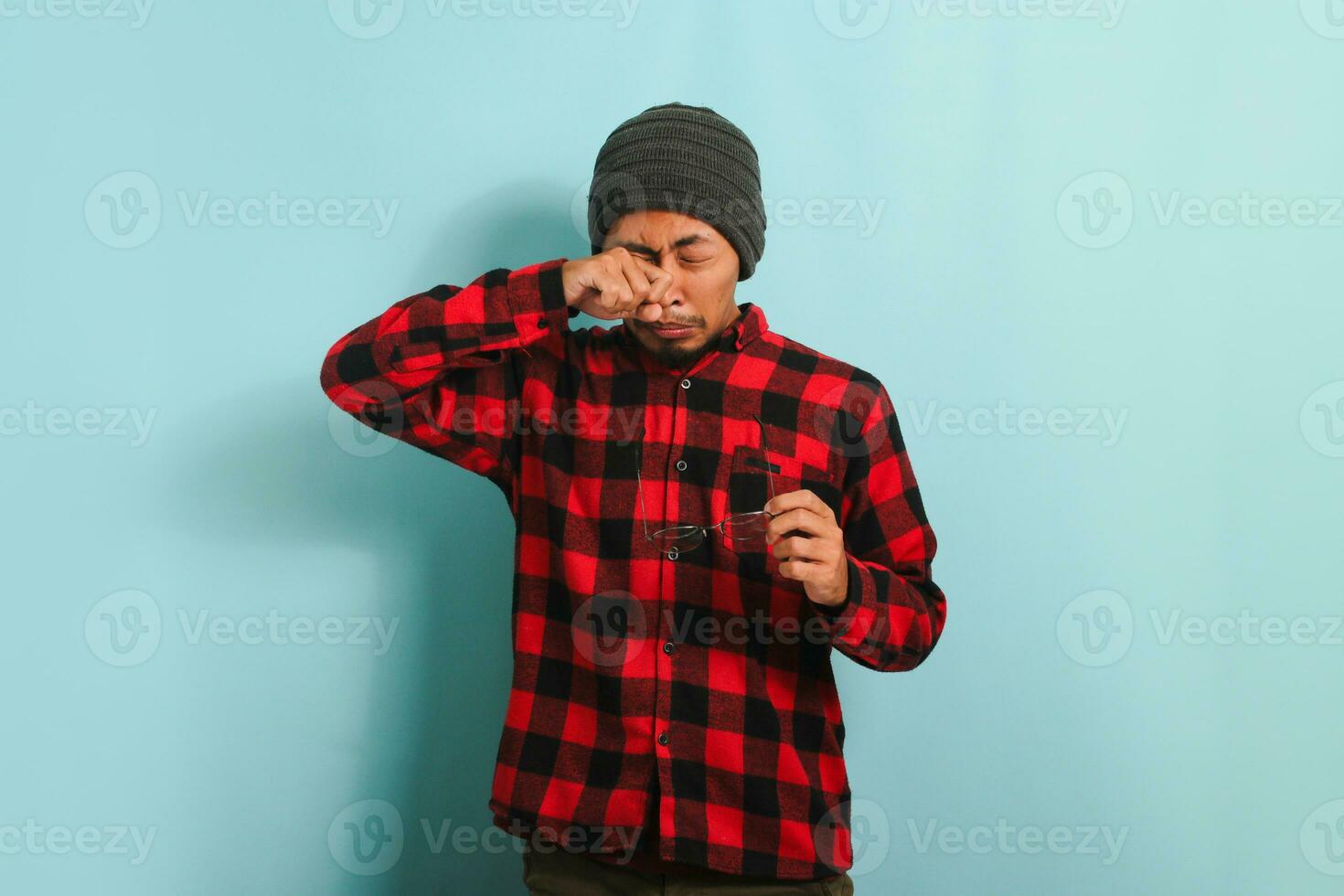 joven asiático hombre con gorro sombrero y rojo tartán franela camisa experimentando incomodidad con contacto lentes, frotamiento su hinchado ojos debido a polvo alergia y picor, aislado en un azul antecedentes foto