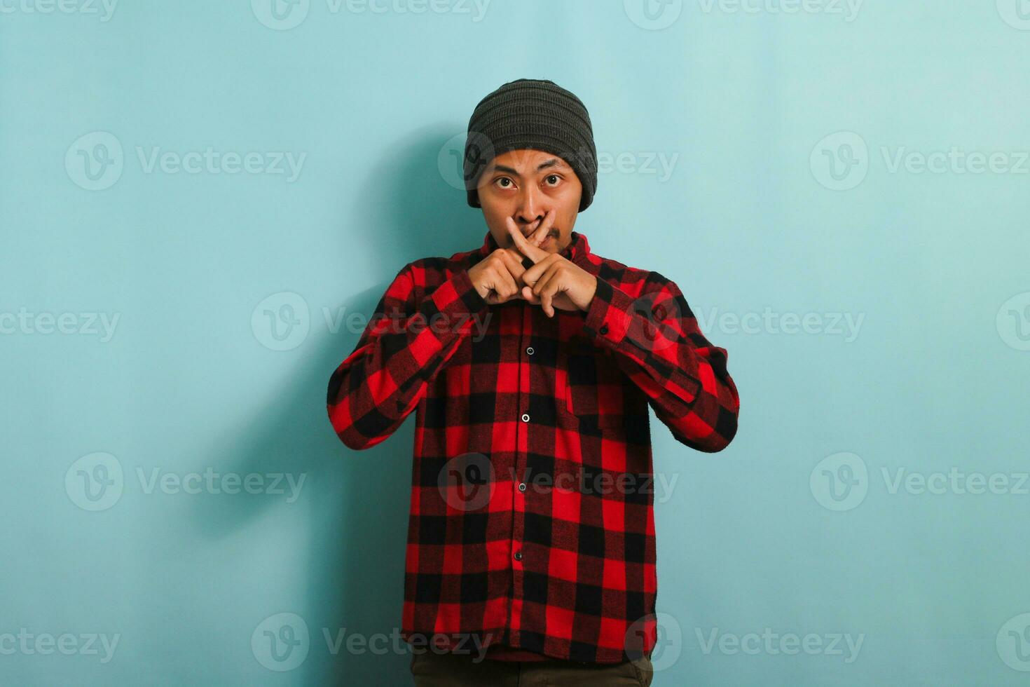 Serious Young Asian man with beanie hat and red plaid flannel shirt makes a crossed fingers gesture, indicating to stop talking or stay mute, isolated on a blue background photo