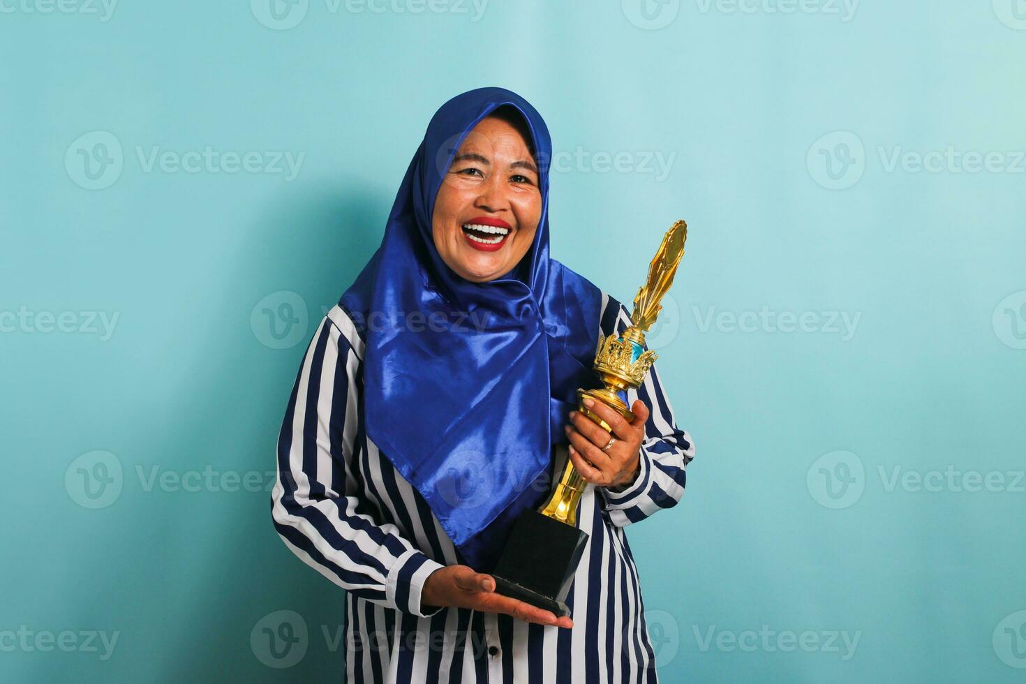 un emocionado de edad mediana asiático mujer de negocios en un azul hijab y un a rayas camisa es sonriente mientras participación un oro trofeo, celebrando su éxito y logro. ella es aislado en un azul antecedentes foto