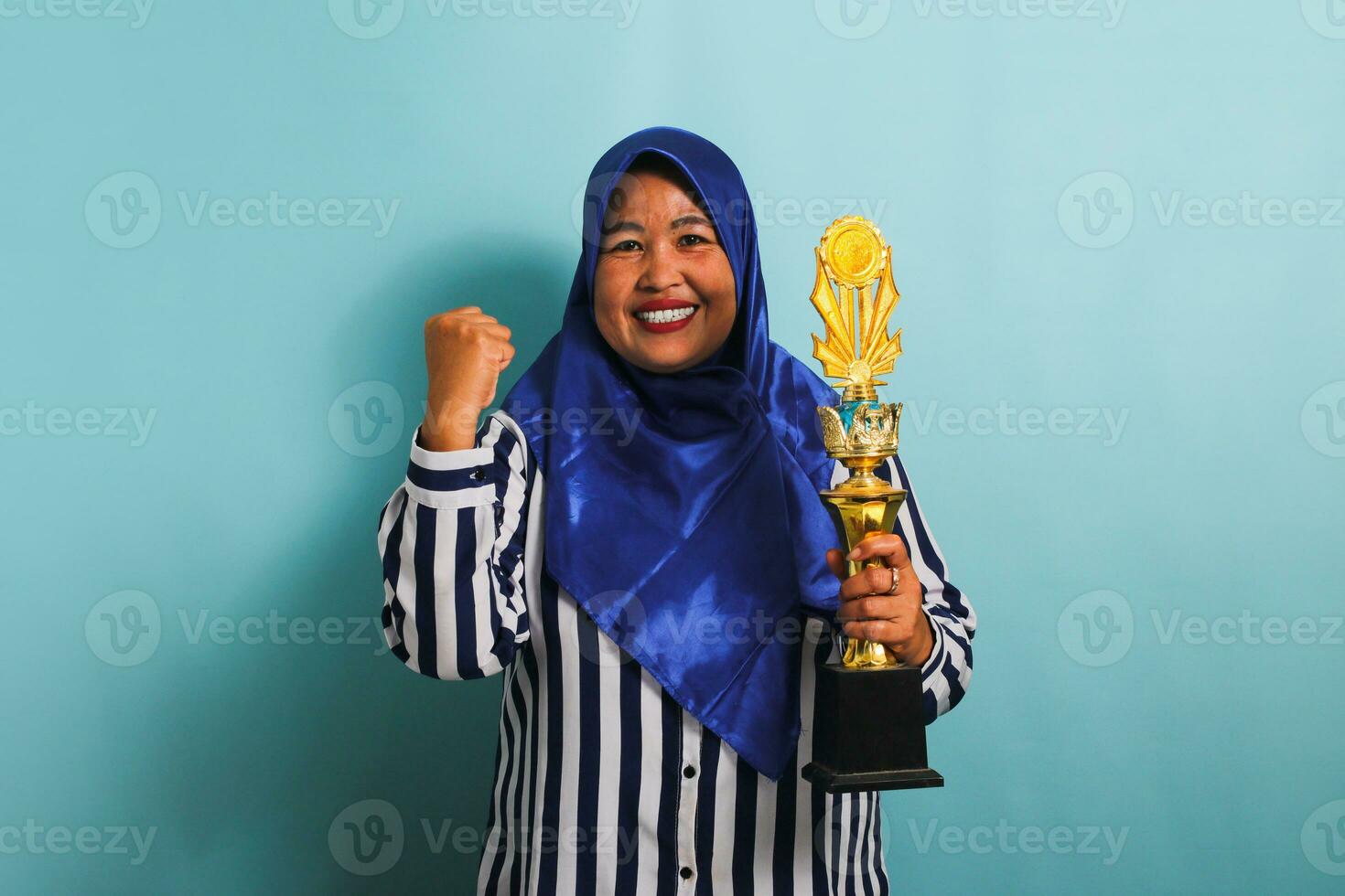 A proud middle-aged Asian businesswoman in a blue hijab and a striped shirt is making a winner gesture while holding a gold trophy, celebrating her success. She is isolated on a blue background. photo