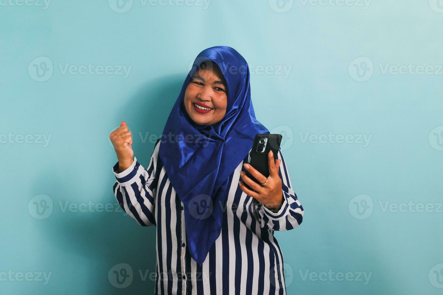 An excited middle-aged Asian woman in a blue hijab and a striped shirt is showing an enthusiastic expression while holding a mobile phone. She is isolated on a blue background photo