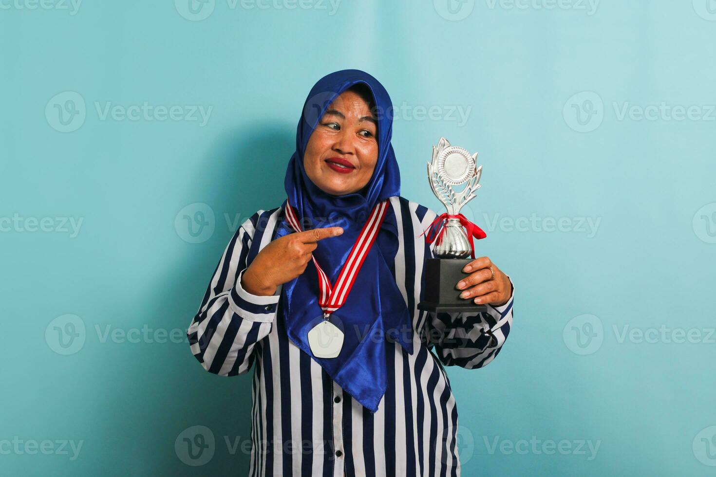 A happy middle-aged Asian businesswoman in a blue hijab, striped shirt, and medal is pointing a silver trophy, celebrating her success and achievement, isolated on a blue background photo