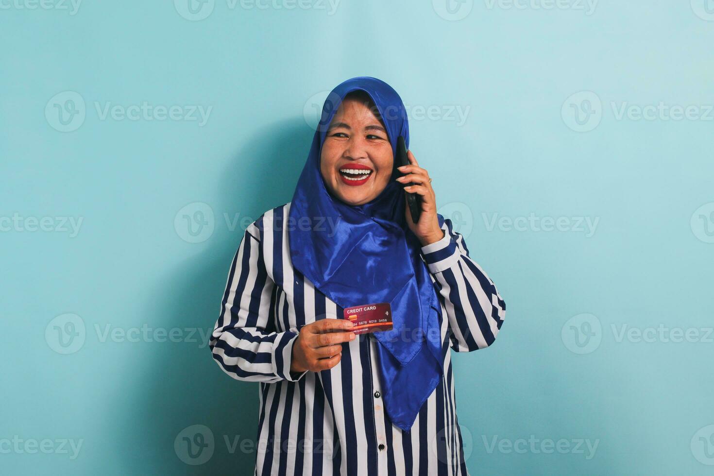An excited middle-aged Asian woman in a blue hijab and a striped shirt is talking on the phone and showing a credit card. She is isolated on a blue background photo