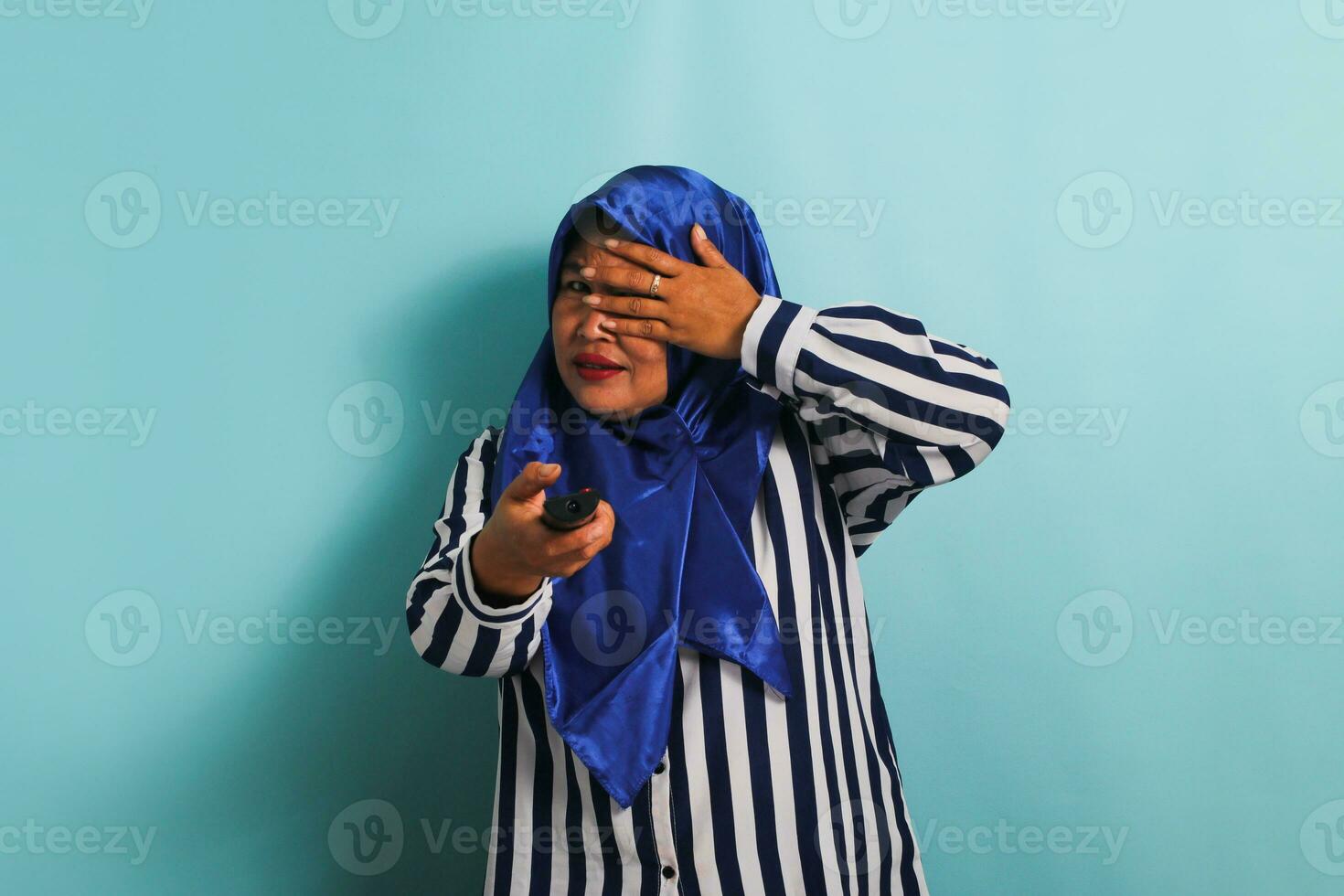 un asustado de edad mediana asiático mujer en un azul hijab y un a rayas camisa es participación un remoto controlar y acecho un horror película en televisor, echar un vistazo mediante su dedos. ella es aislado en un azul antecedentes foto