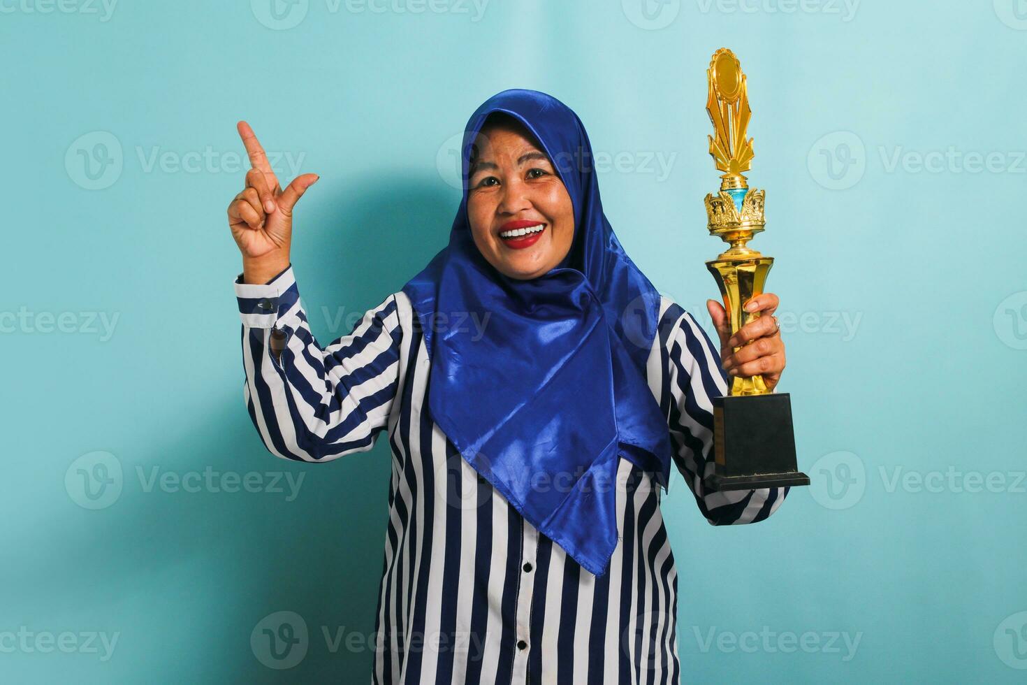 Happy middle-aged Asian businesswoman in blue hijab and striped shirt is holding a gold trophy and pointing up to the copyspace, celebrating her success and achievement, isolated on blue background photo