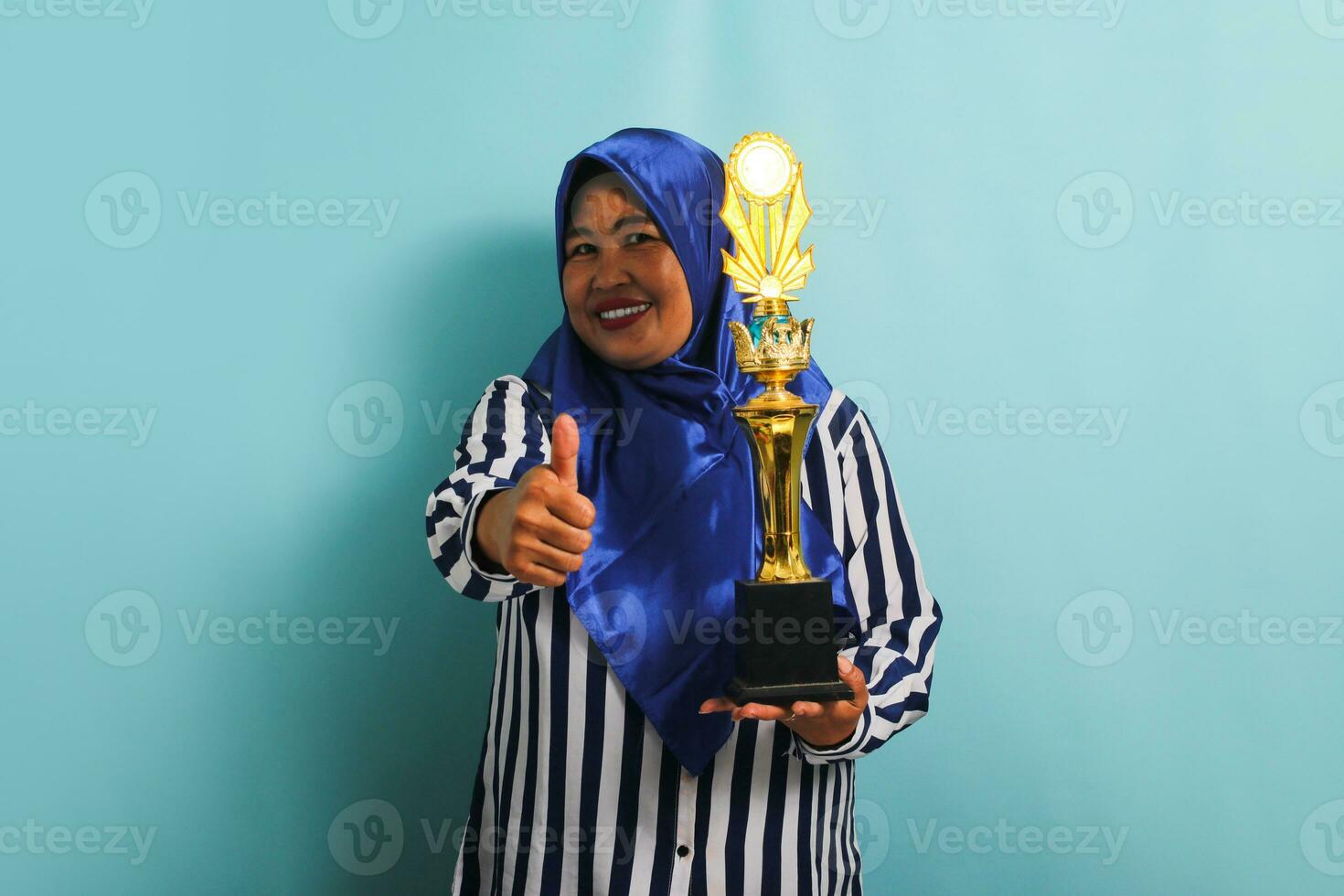 A proud middle-aged Asian businesswoman in a blue hijab and a striped shirt is making a thumb up gesture while holding a gold trophy, celebrating her success. She is isolated on a blue background. photo