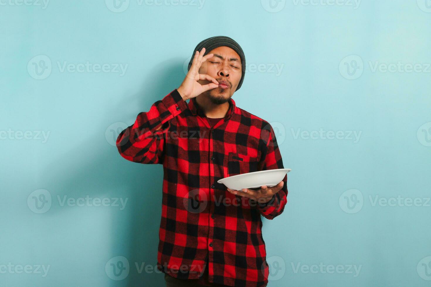 joven asiático hombre es haciendo delicioso mano gesto mientras participación lámina, aislado en azul antecedentes foto