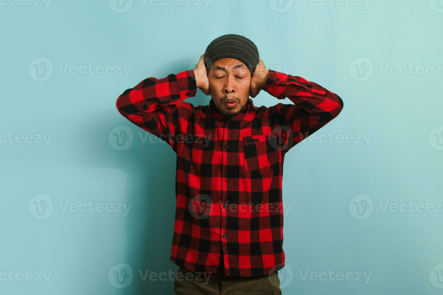 A young Asian man with a beanie hat and a red plaid flannel shirt is suffering from a headache, feeling desperate and stressed due to the pain and migraine, while standing against a blue background photo