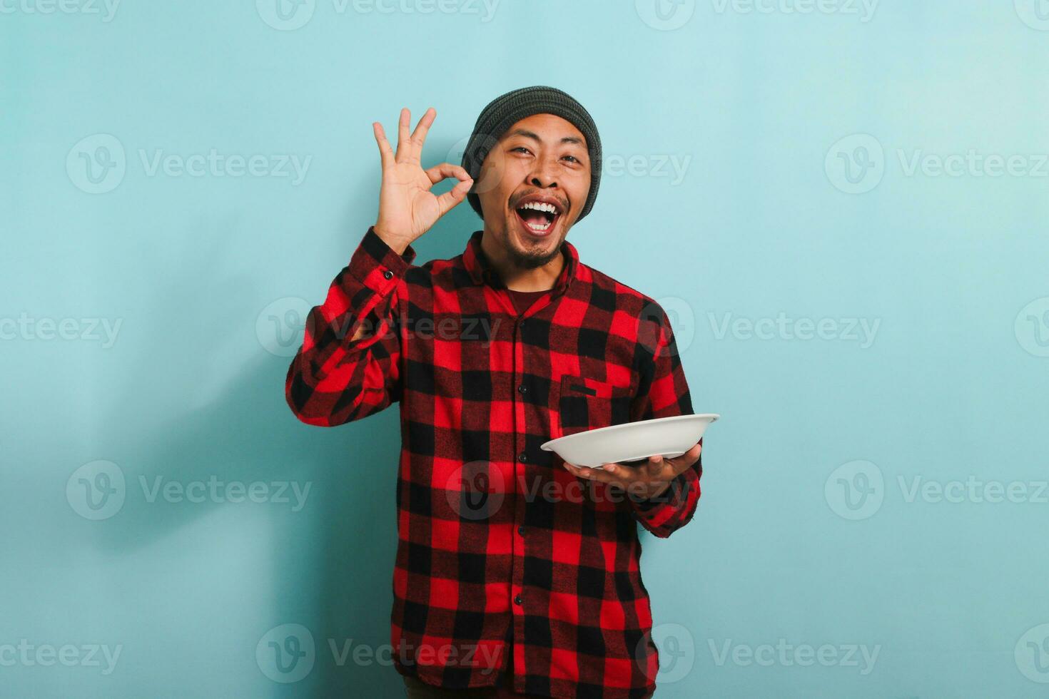 Satisfied Young Asian man with beanie hat and red plaid flannel shirt is making delicious hand gesture to express how good the food is while holding an empty white plate, isolated on a blue background photo