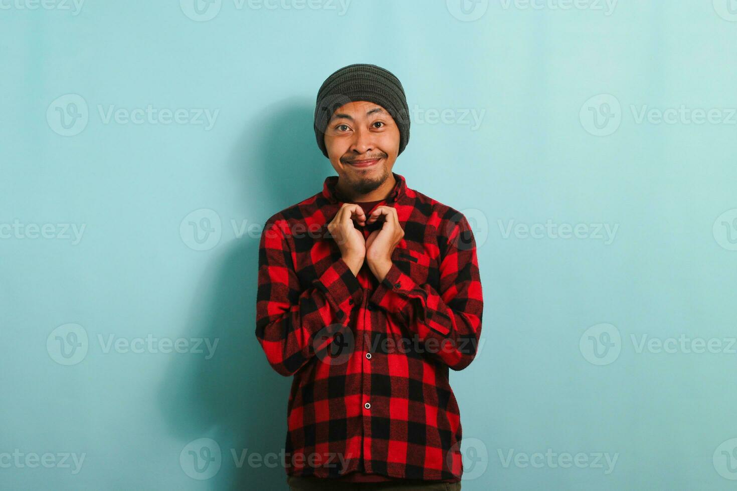 Smiling young Asian man with a beanie hat and a red plaid flannel shirt shows a love gesture with his hand while standing against a blue background photo