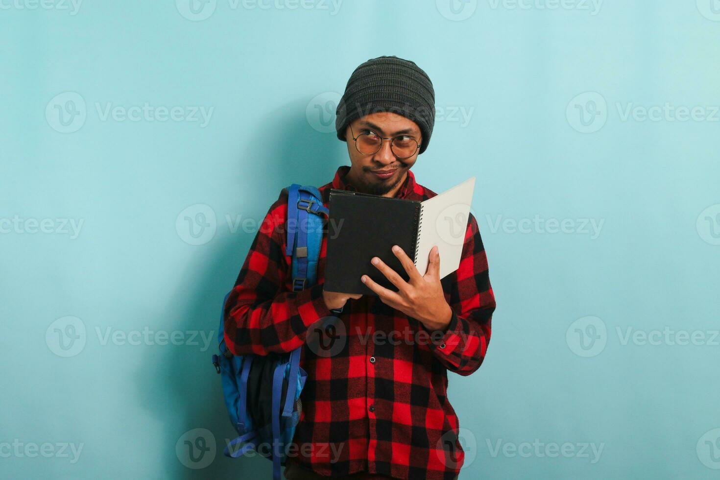 Pensive young Asian student man is thinking while holding book and standing against blue background photo