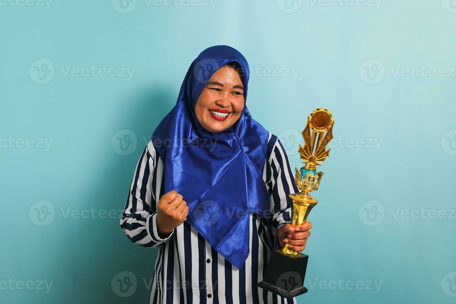 un orgulloso de edad mediana asiático mujer de negocios en un azul hijab y un a rayas camisa es haciendo un ganador gesto mientras participación un oro trofeo, celebrando su éxito. ella es aislado en un azul antecedentes. foto