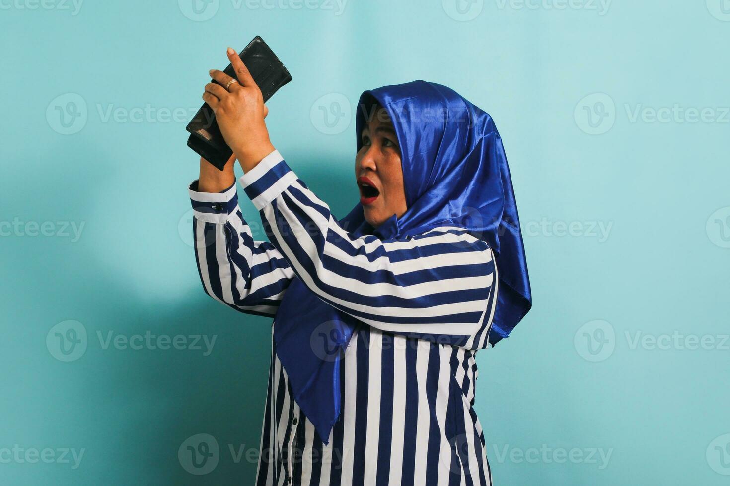 un asombrado de edad mediana asiático mujer en un azul hijab y un a rayas camisa es mirando dentro su vacío billetera con un conmocionado y sorprendido expresión. ella es aislado en un azul antecedentes foto