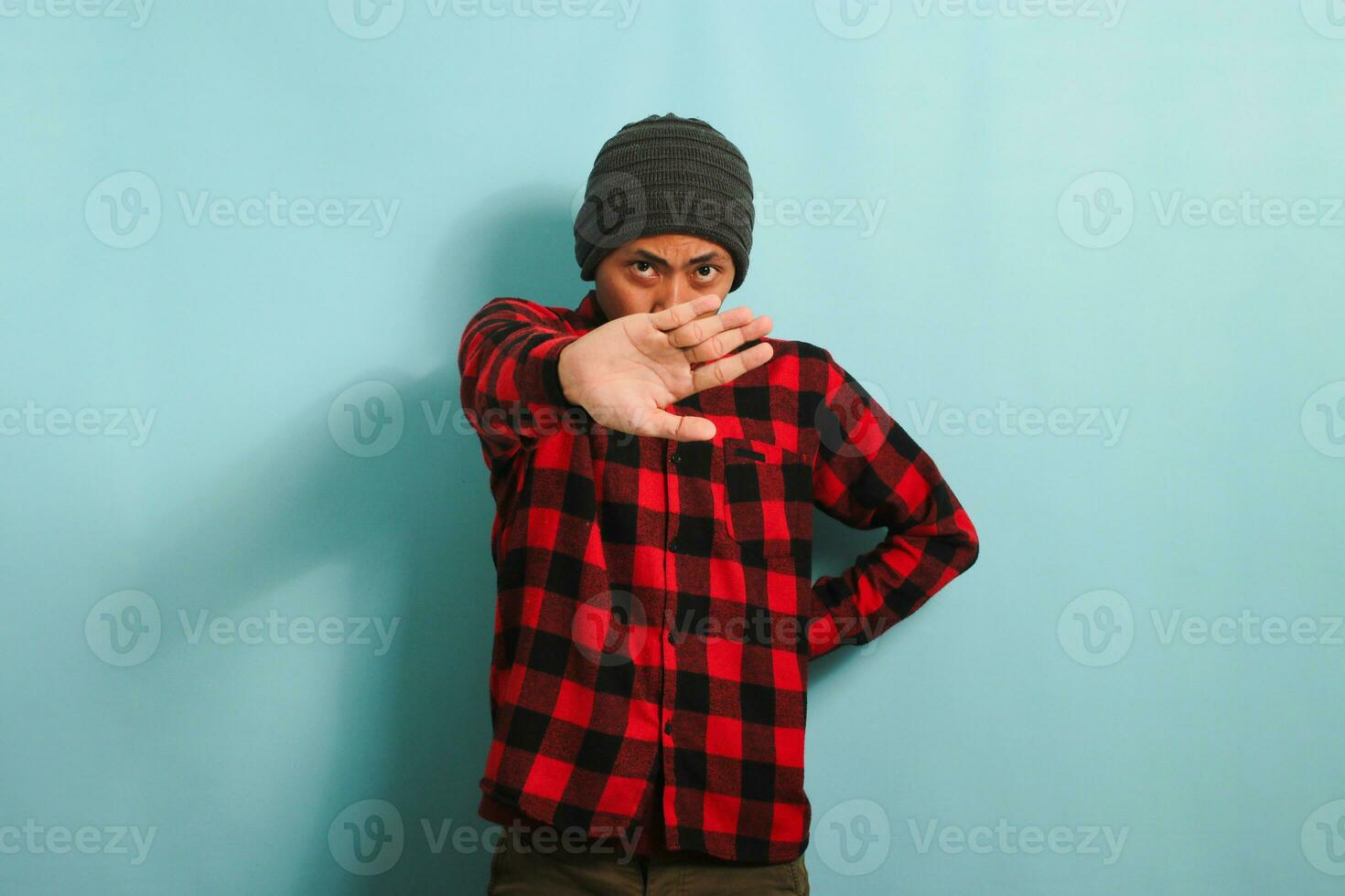Angry young Asian man stands with his arms crossed, stop gesture, isolated on blue background. photo