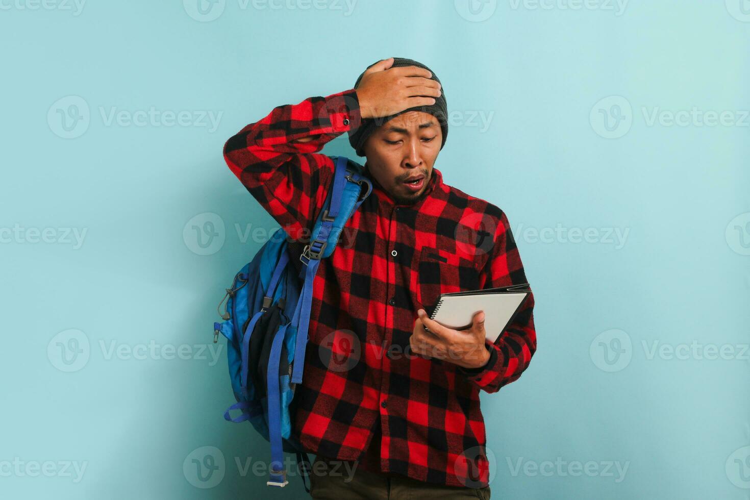 Surprised young Asian student appears unhappy with his hand on his head, isolated on blue background photo