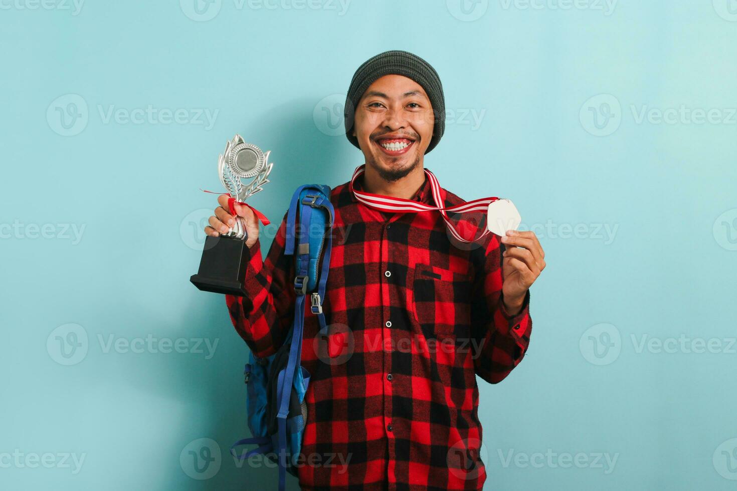 contento joven asiático hombre estudiante demostración un medalla y trofeo, aislado en un azul antecedentes foto