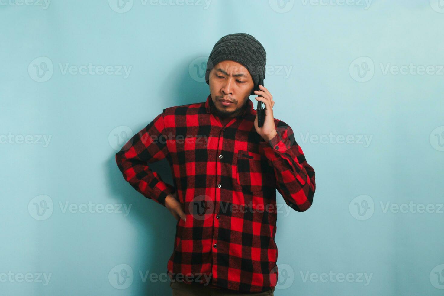 grave joven asiático hombre con un gorro sombrero y un rojo tartán franela camisa tiene un preocupado y infeliz Mira mientras hablando en su móvil teléfono y recepción malo noticias, aislado en un azul antecedentes foto