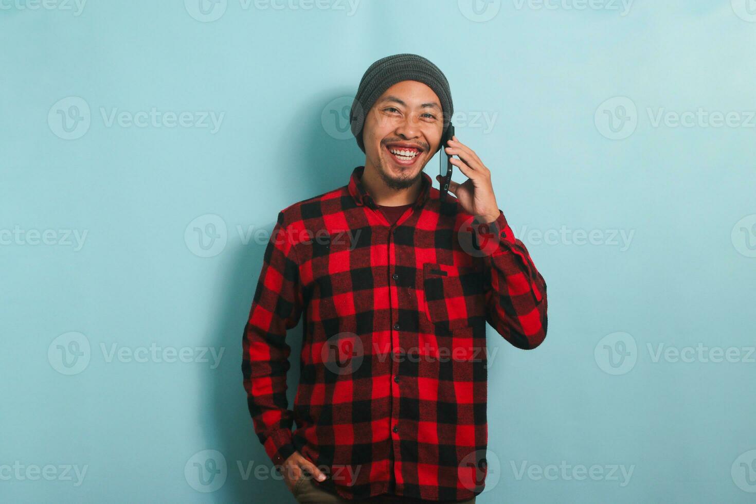 Confident Young Asian man with a beanie hat and a red plaid flannel shirt is smiling at the camera while talking on his smartphone, isolated on a blue background photo