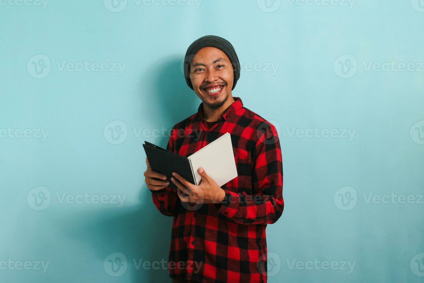 A smiling young Asian man with a beanie hat and a red plaid flannel shirt is holding a book while standing against a blue background photo