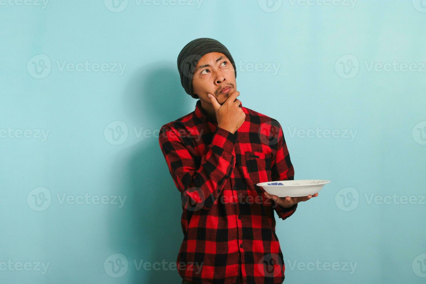 pensativo joven asiático hombre pensando acerca de qué a comer mientras participación lámina, aislado en azul antecedentes foto