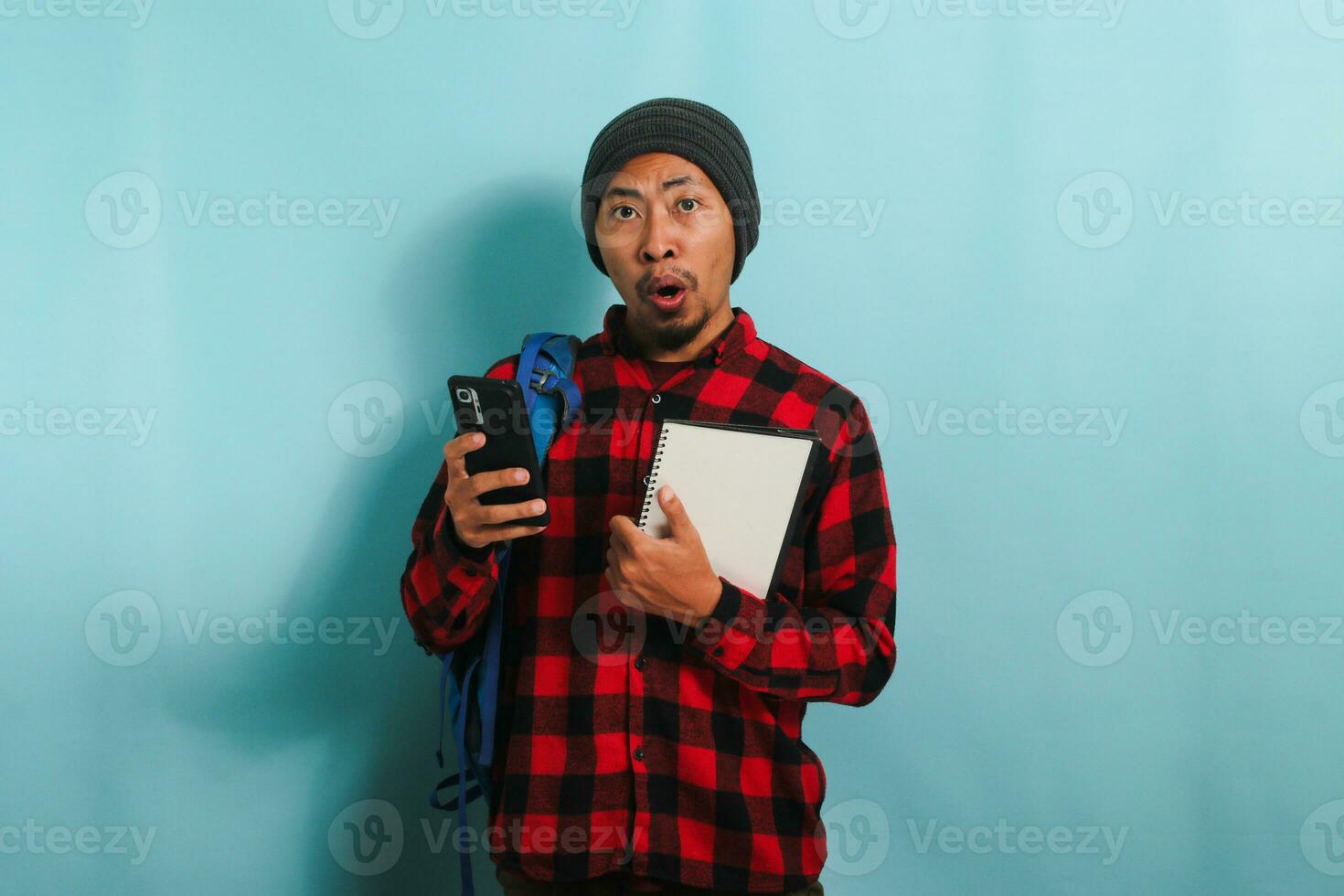 Puzzled young Asian male student, wearing backpack, beanie hat, and red plaid flannel shirt, looks shocked, has forgotten to learn a lecture and is not prepared for exams, isolated on blue background. photo