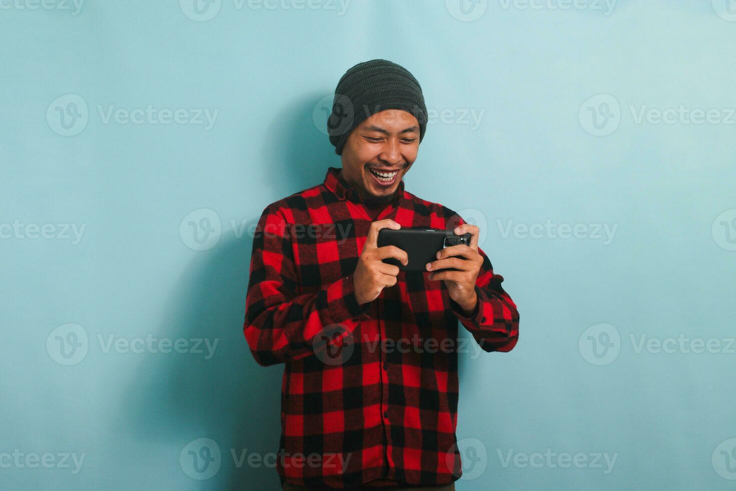 emocionado joven asiático hombre con un gorro sombrero y un rojo tartán franela camisa es jugando un en línea juego en su teléfono inteligente, aislado en un azul antecedentes foto