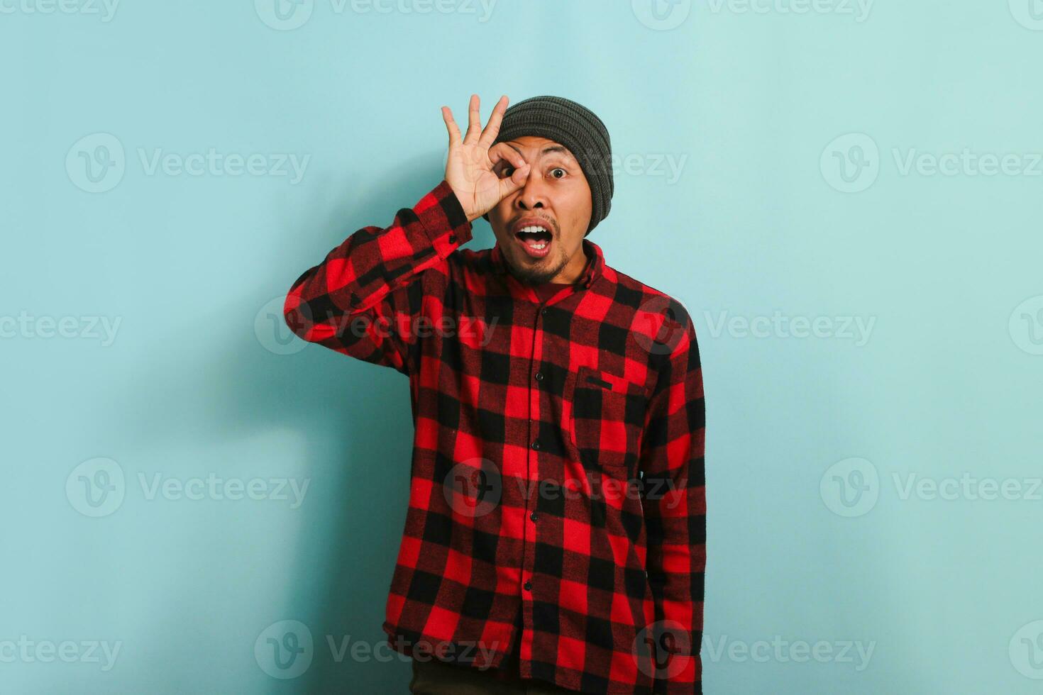Excited young Asian man makes the OK sign gesture like binoculars, isolated on blue background photo